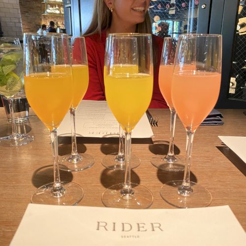 The image shows a woman sitting at a table with five glasses of colorful drinks and a menu from a place named "Rider."