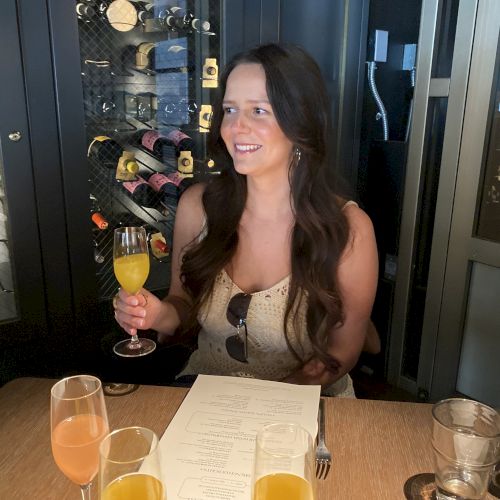 A woman is seated at a table with glasses of orange and peach drinks; behind her is a display of wine bottles. She is smiling while holding a glass.
