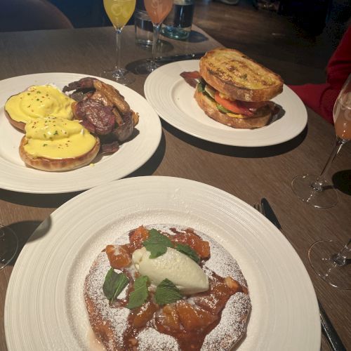 The image shows a table with three plates of food, including eggs Benedict, a sandwich, and a dessert, along with glasses of beverages.