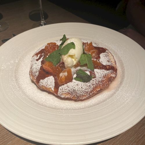 A dessert dish featuring a pastry topped with whipped cream, mint leaves, and caramelized fruits, dusted with powdered sugar, on a white plate.