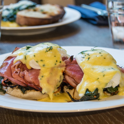 A plate of eggs Benedict with hollandaise sauce, ham, and spinach on toasted English muffins, with another dish in the background.