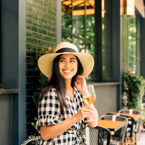 A smiling woman in a wide-brimmed hat holds a glass of wine, standing outside a café with a checkered dress, surrounded by greenery and empty tables.