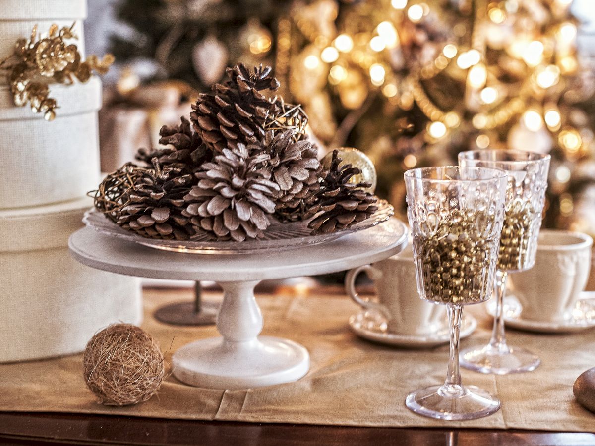 A festive table with pinecones on a stand, decorative glasses, teacups, and a lit Christmas tree in the background.