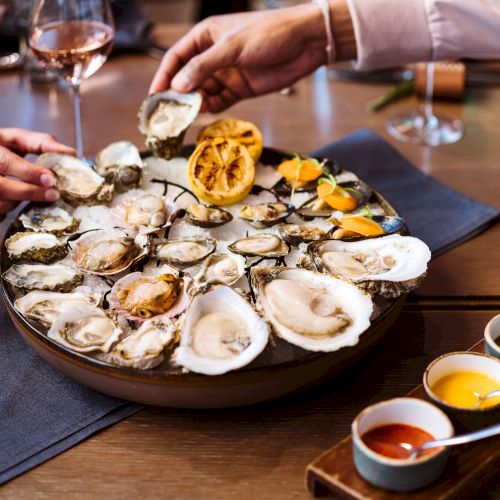 A platter filled with oysters is being shared among people, accompanied by lemon, sauces, and glasses of wine on a dining table.