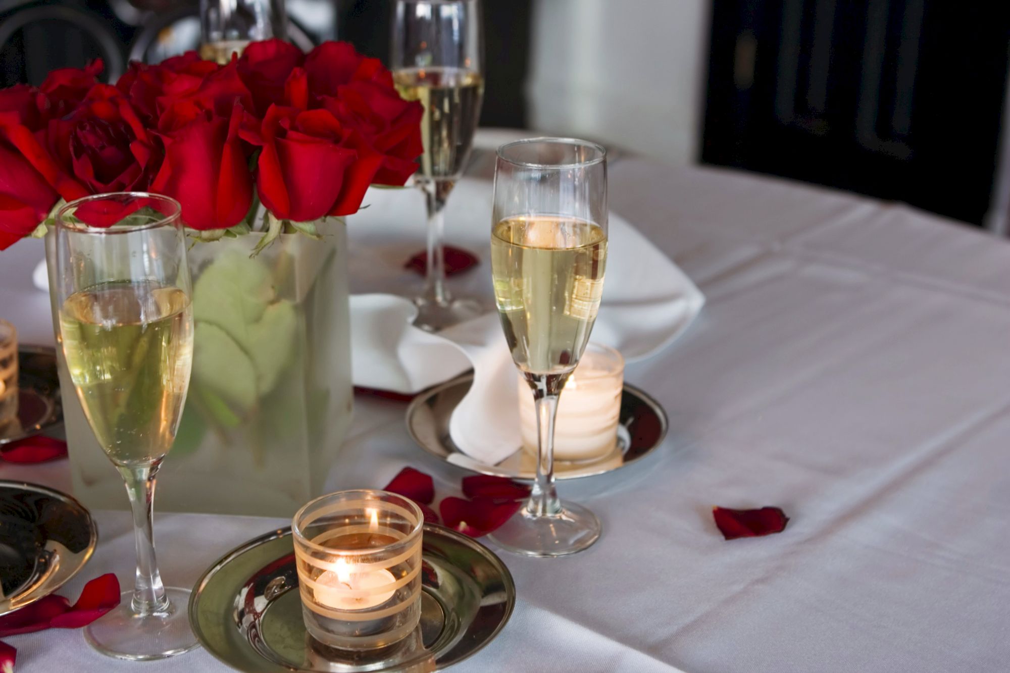 The image shows a table set with champagne glasses, red roses, and lit candles on silver trays, creating an elegant, romantic ambiance.