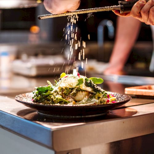 A chef is grating cheese over a dish garnished with greens, showcasing a professional kitchen setting.