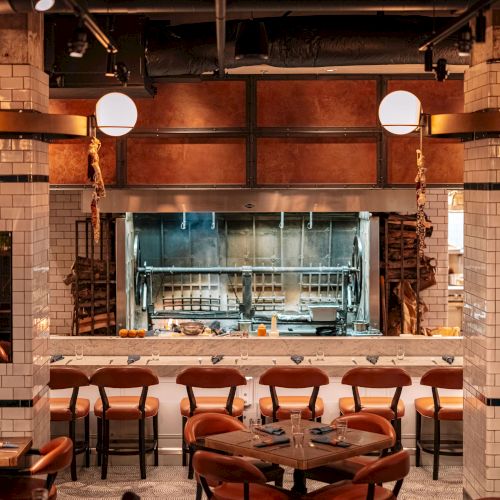 A stylish restaurant interior with a rotisserie, elegant lighting, and neatly arranged tables and chairs for dining.