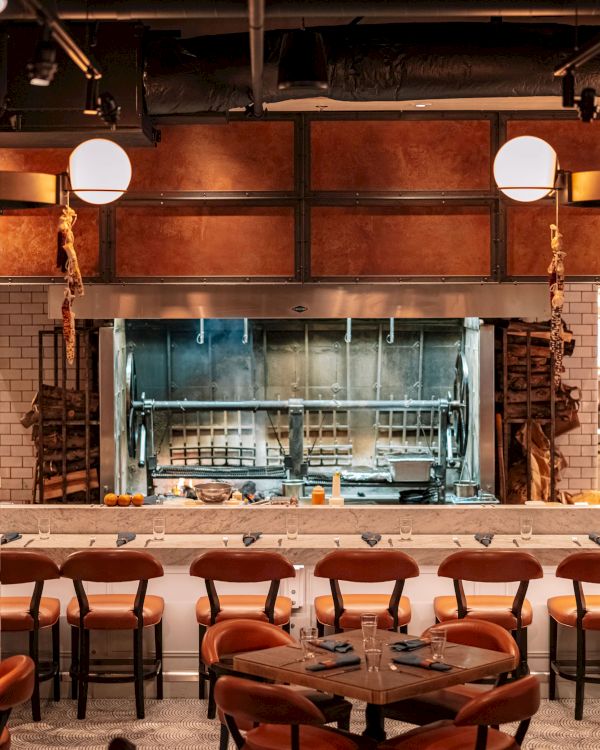 A stylish restaurant interior with a rotisserie, elegant lighting, and neatly arranged tables and chairs for dining.