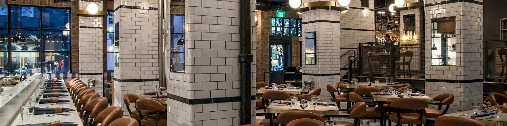 A modern restaurant dining room with set tables, tan chairs, white-tile pillars, and warm pendant lights amid exposed ductwork.