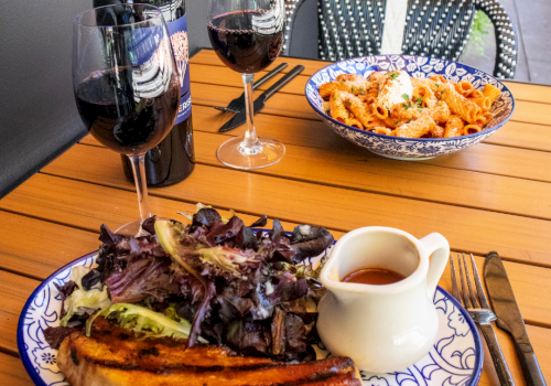 A sunny outdoor table with red wine, two glasses, pasta and salad plates with sauce for dipping.